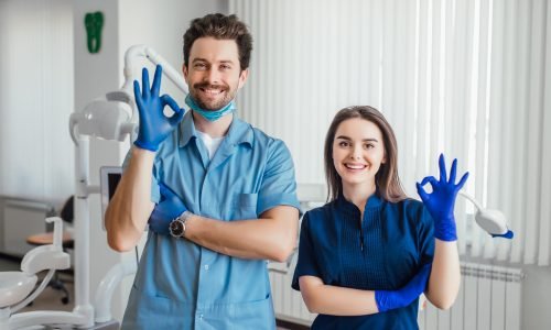 Photo of smiling dentist standing with arms crossed with her colleague, showing okay sign.